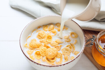 Pouring of milk into bowl with cornflakes on light wooden background, closeup