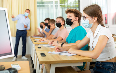 Teenage students in protective mask studying in classroom with teacher, writing lectures in workbooks