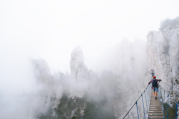 A young man in mountaineering equipment walks uphill on a suspension bridge in the fog. VIA Feratta - Heavenly trail. Suspension bridge on Mount Ai-Petri, Yalta, Crimea. Sea coast in Crimea.  - obrazy, fototapety, plakaty