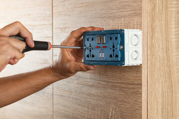 Electrician is using a screwdriver to install a power outlet in to a plastic box on a wooden wall.