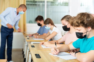 Fototapeta premium Pupils in face masks sitting in class and listening carefully to male teacher.