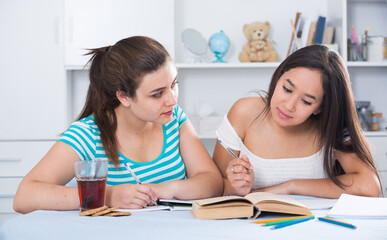 Two girls classmates sitting at table at home and doing homework together
