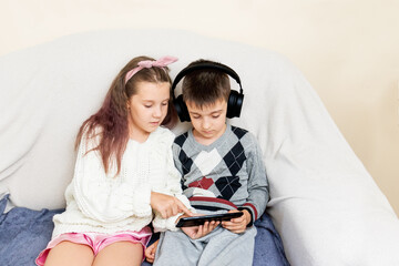 brother and sister are sitting on the couch in the living room and watching a tablet at home. boy in headphones and girl showing finger to tablet.  
the concept of friendship between brother and siste