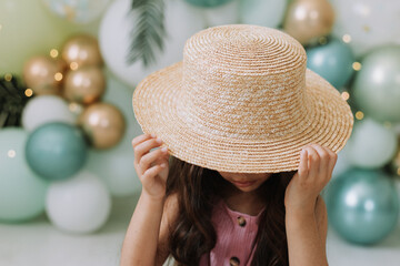 Closeup portrait of a beautiful smiling little dark-skinned girl with brown eyes and dark hair in a straw hat against the background of balloons. Concept of spring summer youth beauty. Beautiful face