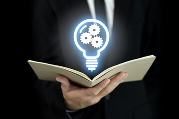 Smart businessman in black business suit holding a book on hand with illuminated lightbulb and gears icon.