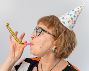 Portrait of a smiling elderly woman in a festive cap holding a whistle tongue on a white background