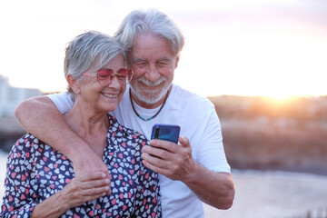 Youthful senior couple embracing in outdoor at sea at sunset using mobile phone. Caucasian retired enjoying relax and happy lifestyle