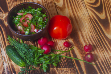 Vegetable salad in a clay plate of tomatoes, cucumbers, radishes, dill, onions, parsley on a wooden background