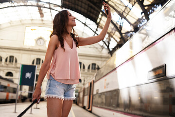 Young woman waiting for the train standing at the railway station. Beautiful girl taking selfie photo.