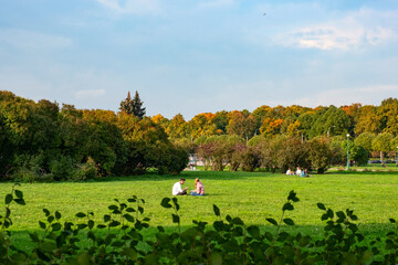 A couple of young people are sitting on the green grass on great lawn grass meadow in autumn fall...