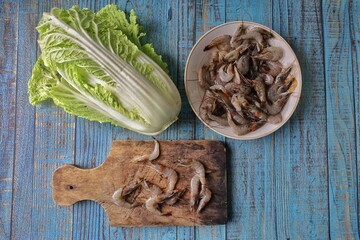 a bunch of raw prawns on a wooden placemat and a white plate with mustard greens on a blue painted wooden background