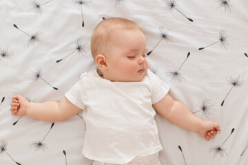 Indoor shot of cute charming adorable baby sleeping on bed, wearing white t shirt, keeping eyes...
