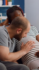 Interracial couple expecting child in living room. Caucasian husband talking to baby and touching belly while african american wife relaxing on couch at home. Mixed race partners