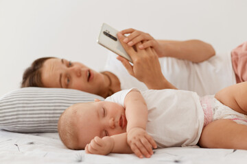 Obraz na płótnie Canvas Portrait of surprised female wearing white t shirt and holding smart phone, looking at smartphone screen with open mouth and astonishment, lying with sleeping baby daughter.