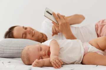 Obraz na płótnie Canvas Happy smiling with with pleasant appearance wearing white casual style t shirt using cell phone, looking at device screen with positive emotions, lying on bed with her little sleeping daughter.
