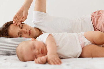Obraz na płótnie Canvas Portrait of dark haired female wearing white casual t shirt lying on bed with little baby daughter, posing indoor, woman looking at her infant girl with tired expression.