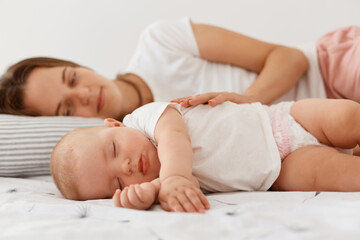 Obraz na płótnie Canvas Indoor shot of smiling female lying with sleeping girl daughter, resting together with her lovely baby, woman wearing white casual t shirt looking at infant girl with positive emotions.