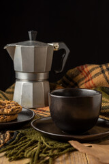 Close-up of black coffee cup with its plate, coffee pot and cookies, on green plaid blanket, wooden table, black background, vertical