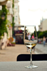 Glass of white wine close up. Alcoholic drink on a wooden table on a background of the city. Transparent wine on the summer terrace of the restaurant. Blurred people in the background.