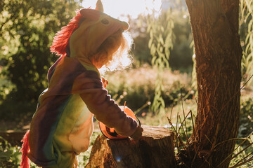 little girl in a rainbow unicorn Halloween costume with a pumpkin basket for sweets is sitting on a stump at a forest sunset. a fabulous wonderful magical forest. space for text. High quality photo