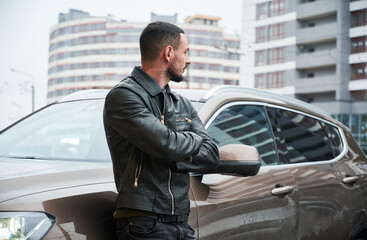 Young stylish man standing on the street with his crossed arms, leaning against luxury car and...