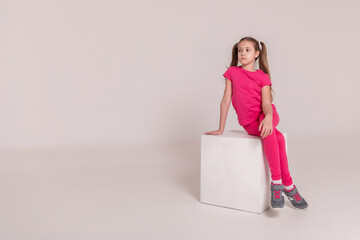 beautiful girl in a sports pink T-shirt and pants on a white isolated background. child sitting on a white cube