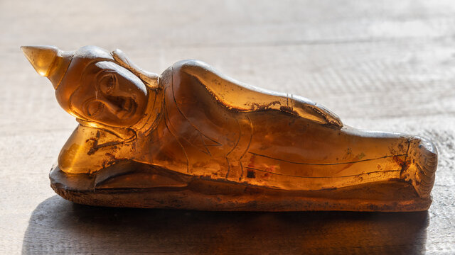 Closeup view of beautiful backlit translucent brown crystal reclining buddha statue on bright wooden background - Powered by Adobe