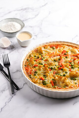 Baked french vegetable pie quiche filled with carrots, peas and cheese on a marble surface, cracked egg shell, flour, cream