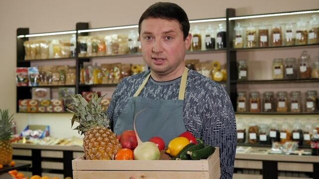 Portrait of a young male salesman in an apron with a box of fresh fruit in his hands, in the background of a shelf with organic products. A healthy food store.