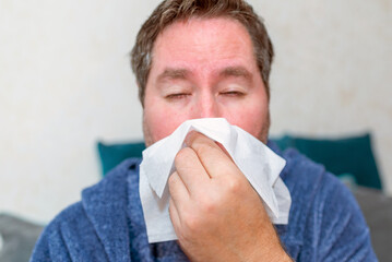 healthcare, people and medicine concept - ill man blowing nose at home. sick male at home have health problems, get flu or fever symptoms, tired guy suffer from sickness sneezing holding tissue.