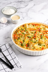 Baked french vegetable pie quiche filled with carrots, peas and cheese on a marble surface, cracked egg shell, flour, cream