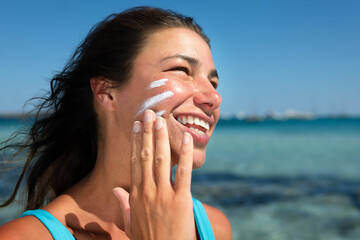 Close up of happy smiling young woman is applying sunscreen or sun tanning lotion on face to take...