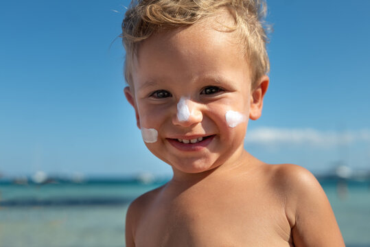 Cute happy little toddler boy is smiling in camera with protective sunscreen or sunblock lotion on his face applied by his mother to take care of skin on seaside beach during family holidays vacation.