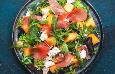Fresh salad with cantaloupe melon, prosciutto, soft cheese and arugula on blue background, top view, copy space