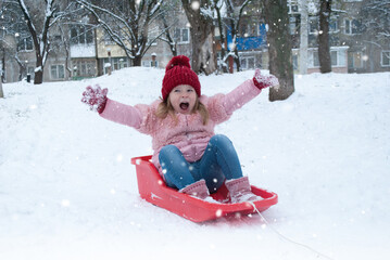 little cute girl on a red sled on white snow in winter on New Year's Day and Christmas raising her hands up to rejoice in the red hat of Santa with a place for the text