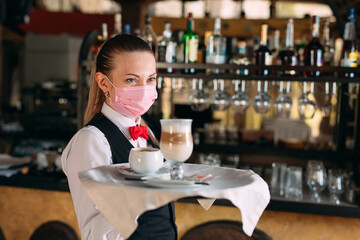 A female Waiter of European appearance in a medical mask serves Latte coffee.