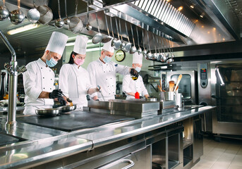 Chefs in protective masks and gloves prepare food in the kitchen of a restaurant or hotel.