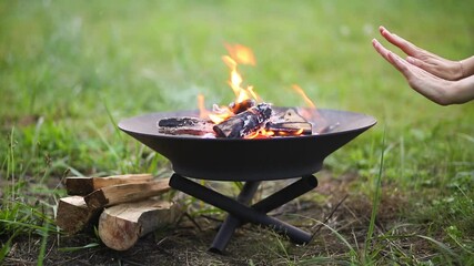 The flame of the fire burns in a metal fire bowl - warm your hands by the fire, stir the firewood with a stick. A hearth with firewood for safe campfire outdoors. Warmth, comfort for gatherings