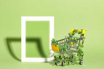 Shopping cart entwined with plants next to empty mock up frame on green background. Conscious...