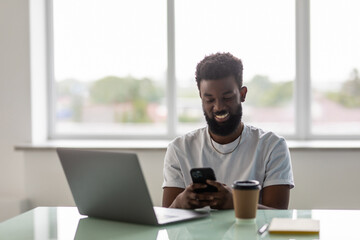 Smiling young african American man sit relax on couch at home browse wireless internet on modern cellphone. Happy male in spectacles look at screen message text use smartphone