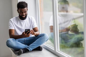 Handsome african man sits home on windowsill, reads text messages, sends email, uses his phone and fast internet connection.