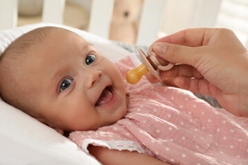Mother giving pacifier to cute little baby in soft crib, closeup
