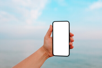 Hand showing a blank smart phone on the beach with the sea in the background. White screen mock up