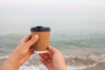 Coffee at the sea. Young woman holds a disposable cup of coffee on on a sunny morning at the beach. - Powered by Adobe