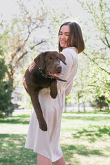Smiling young woman is holding her puppy of chocolate Labrador retriever in arms. Love for pets and friendship concept.