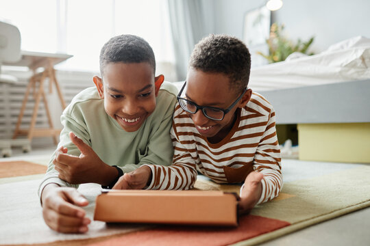 Portrait of two teenage African-American boys using digital tablet together while lying on floor in cozy room, copy space