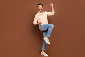 Fototapeta na wymiar Photo of cheerful astonished guy celebrate victory wear beige shirt jeans shoes isolated brown color background
