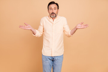 Photo of mature brunet man shrug shoulders wear peach shirt jeans isolated on brown color background