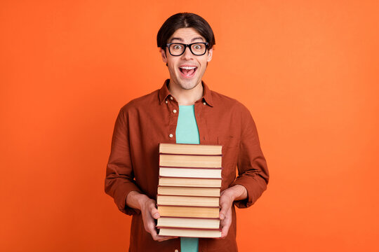 Portrait of charismatic positive smiling college student in glasses hold pile books studying isolated on orange color background