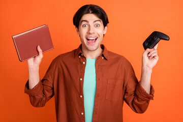 Portrait of young smiling excited scream shout hold playstation and book alternative choice isolated on orange color background
